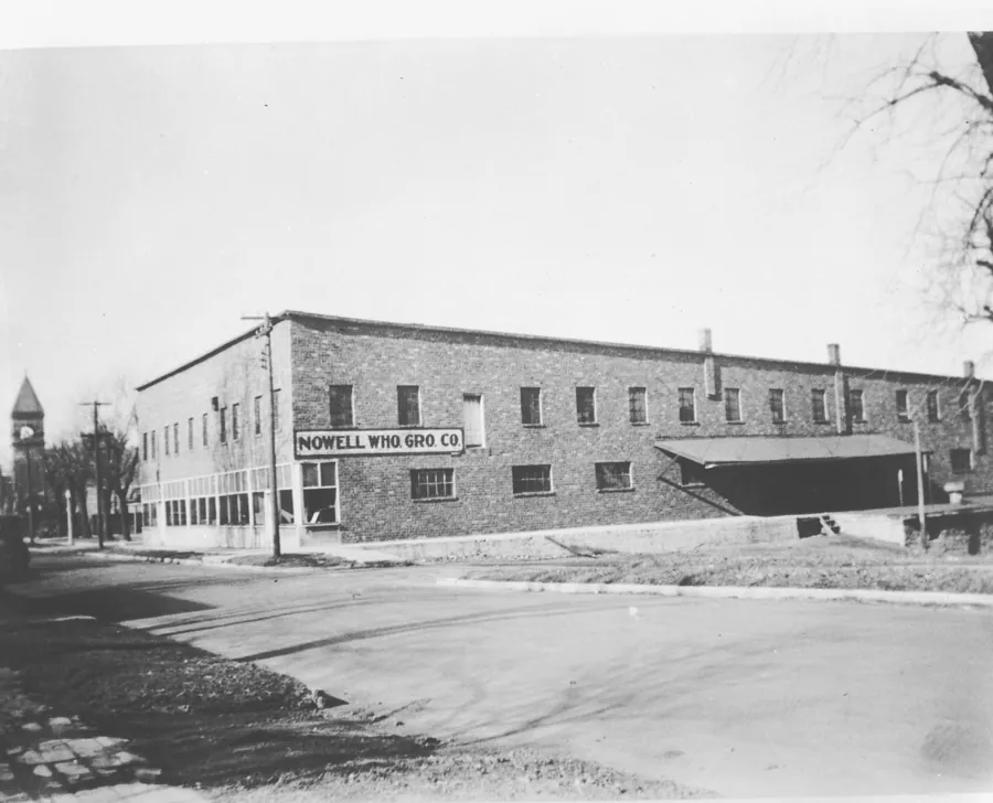 Black-and-white photo of the two-story brick warehouse with a sign reading Nowell Who. Gro. Co., with a loading dock at right and a clock tower in the distance