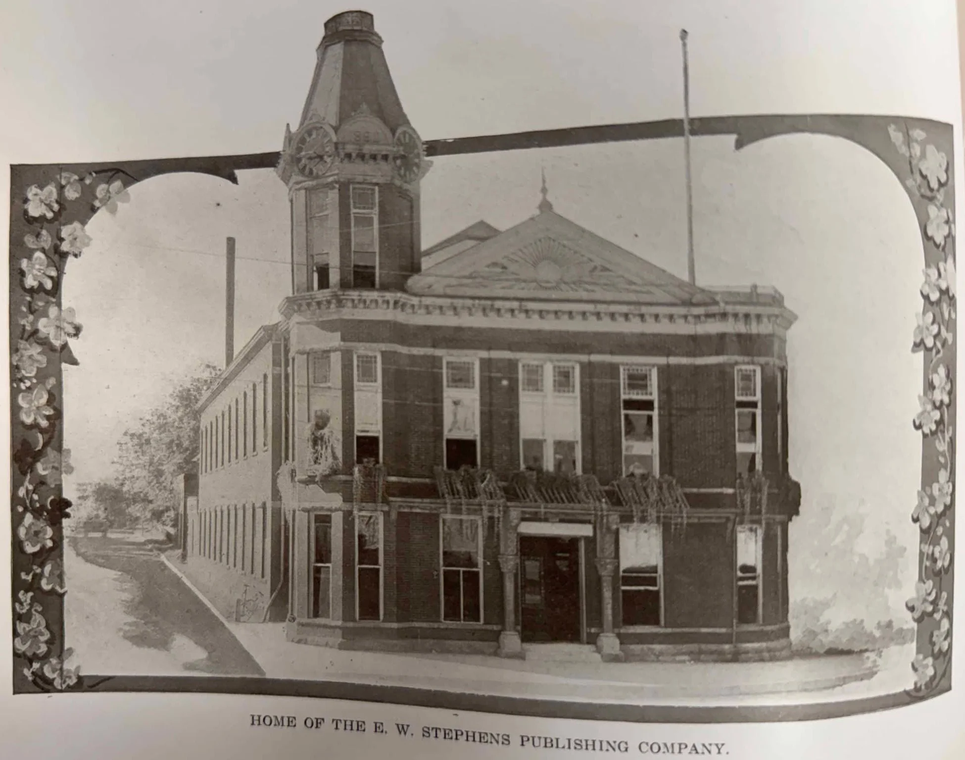 Historic printed photograph of a two-story brick building with a corner clock tower, framed by a flowered border and captioned Home of the E. W. Stephens Publishing Company
