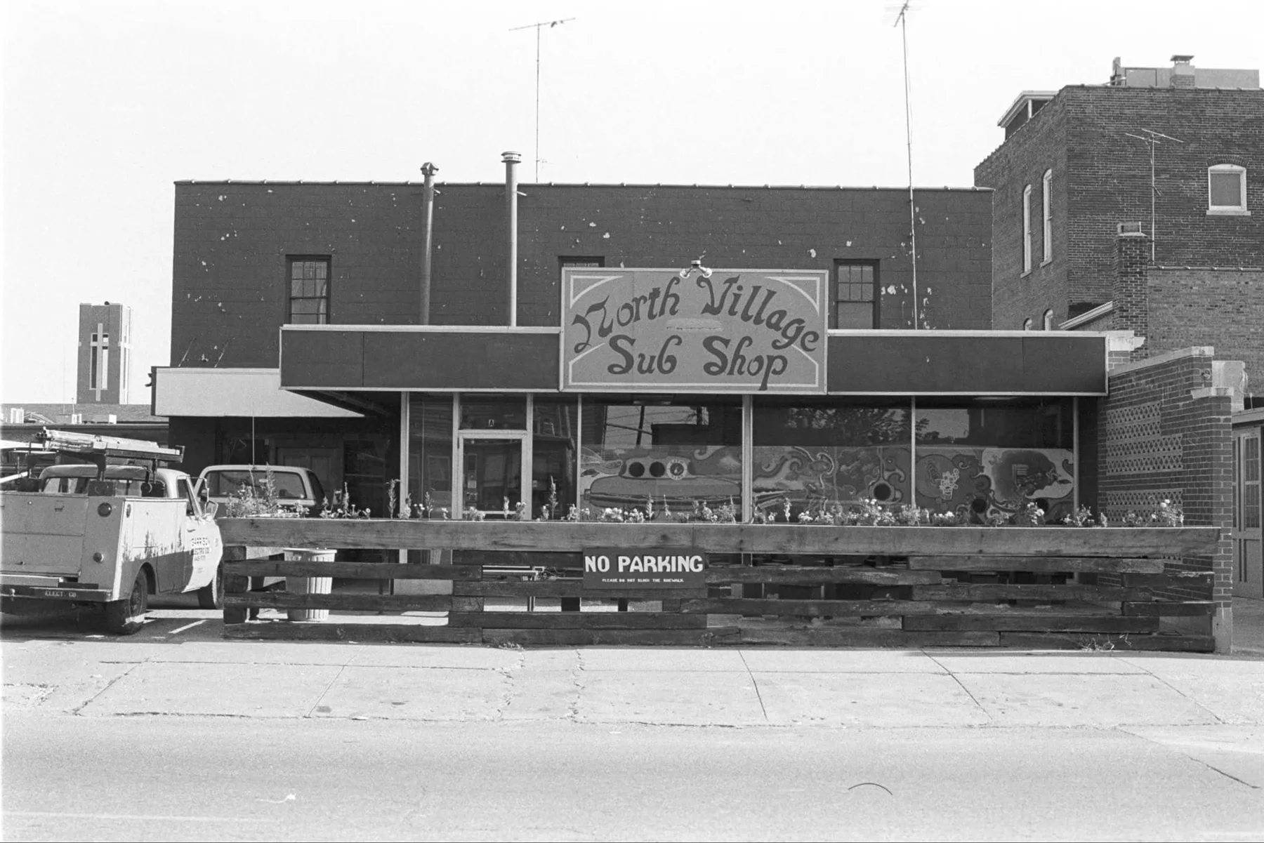 Black-and-white photo of the North Village Sub Shop, with its hand-lettered sign, painted window murals and a railroad-tie patio along the sidewalk